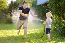 Funny Little Boy With His Father Playing With Garden Hose In Sunny Backyard. Preschooler Child Having Fun With Spray Of Water.