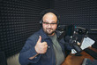 © DedMityay - Funny happy radio presenter or host in radio station studio looks at camera and shows thumbs up gesture, portrait of cheerful working man