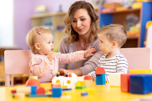 Babies With Mentor In Kindergarten. Kids Toddlers In Nursery School. Little Girl And Boy Preschoolers Playing With Teacher.