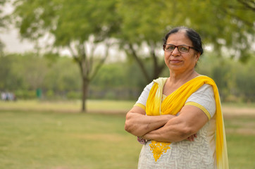 Smart senior north Indian woman standing, posing for the camera with hands crossed in a park wearing yellow salwar kameez in summers in Delhi, India