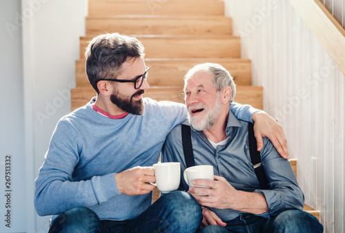 Fotomural An adult hipster son and senior father sitting on stairs indoors at home, talking