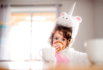 Wall Mural - A portrait of small girl in unicorn mask sitting at the table at home.