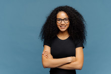 Waist Up Shot Of Smiling Afro American Woman Has Arms Folded, Wears Spectacles And Casual Black T Shirt, Enjoys Nice Conversation With Interlocutor, Poses Over Blue Studio Wall With Blank Space