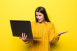 Young pretty woman with shocked face holding laptop wearing in yellow sweater isolated on yellow background