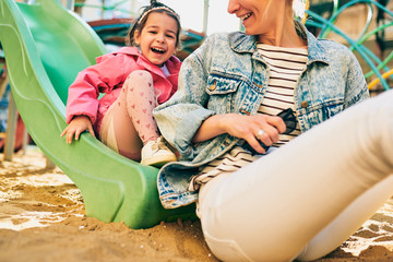 Horizontal outdoor image of happy cute little girl smiling and looking to her beautiful mother spending time together at playground. Young woman and daughter feeling happy, loving each other outside.