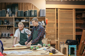 Two woodworker craftsmen are examining the drawings of the product of a new project while standing in their carpentry against the background of shelves with tools. Concept of hard work