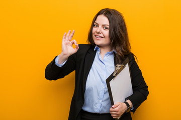 Young curvy plus size business woman holding a clipboard cheerful and confident showing ok gesture.