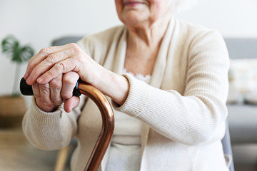 Wall Mural - Elderly woman sitting in nursing home room holding walking quad cane with wrinked hand. Old age senior lady wearing beige cardigan, metal aid stick handle bar close up. Interior background, copy space