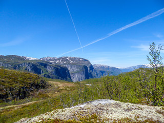 A view on higher parts of Eidfjorden, Norway. Taller mountains have some snow on them. Lush green flora growing on the slopes. Sky is clear blue, with traits of two plains crossing it.