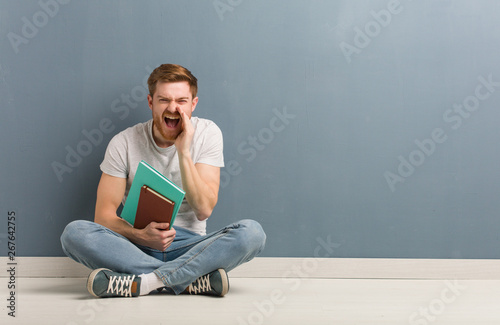 Young redhead student man sitting on the floor shouting something happy to the front Poster Mural XXL