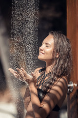 Wall Mural - Wet happy smiling girl enjoying outdoor tropical shower at Maldives. Young woman showering outside by palms at summer vacation. Brunette in black swimwear enjoying natural water on exotic beach.