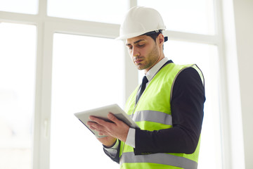 Waist up portrait of  Middle-Eastern engineer wearing hardhat standing against window  holding tablet, copy space