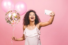 Self Portrait Of Afro American Positive Girl In Dress Having Balloons In Hand Shooting Selfie On Front Camera Isolated On Pink Background.