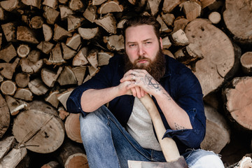 Brutal man with a beard dressed in casual clothes with tattoos on his hands holds the ax sitting near the stacked wood