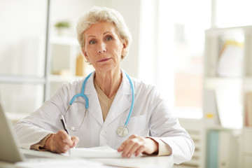 Portrait of senior doctor sitting at desk and looking at camera in office, copy space