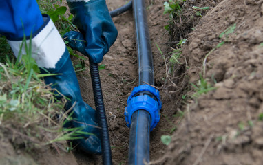 worker in blue uniform puts black water pipes and an electrician in a trench