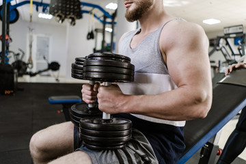 Muscular handsome man training with heavy dumbbell in the gym. Young man with big biceps sits and does workout indoors. Sport and health concept.