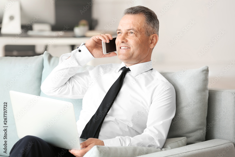 Handsome mature businessman talking by phone while working on laptop in office