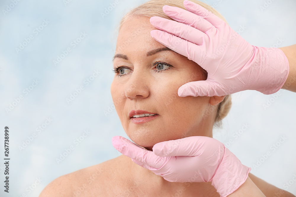 Plastic surgeon touching face of mature woman on light background