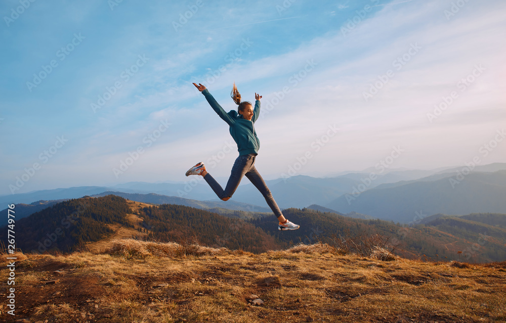 Happy woman hiker jumping on mountain ridge on blue cloudy sky and mountains background. Travel and active lifestyle concept. - obrazy, fototapety, plakaty 