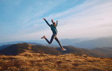Fototapeta  - Happy woman hiker jumping on mountain ridge on blue cloudy sky and mountains background. Travel and active lifestyle concept.