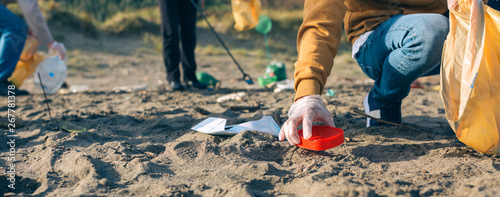 Cuadros en Lienzo Unrecognizable young man picking up trash with group of volunteers on the beach