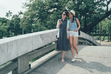 Wall Mural - full length two asian friends having walk on bridge in chinese traditional park in taiwan taichung. young girls communicating and using mobile phones by nature pool. women travelers browse map app