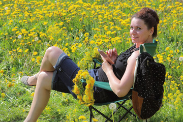 A girl sits in a picnic chair and works with a smartphone. On a spring meadow with bright green young grass and flowering dandelions.  A wreath of dandelions woven. The girl turns around and smiles.