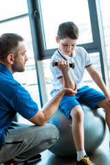 Canvas Print - father helping son on fitness mat exercising with dumbbell at sports center