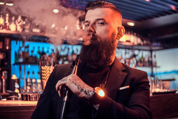 Portrait of tattooed bearded man which is smoking hookah, making nice vapour while sitting near bar counter