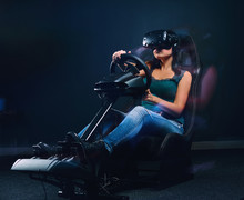 Young Woman Wearing VR Headset Driving On Car Racing Simulator Cockpit With Seat And Wheel.