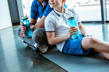 Wall Mural - cropped view of father and son sitting on fitness mat with sport bottles at gym