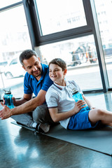 Canvas Print - happy father and son sitting on fitness mat with sport bottles at gym