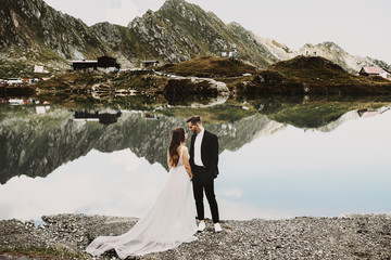 Wall Mural - Full length portrait of a amazing couple in their wedding day near a lake in the mountains looking to each other smiling.