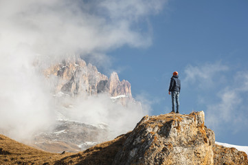 A traveler is a man with a beard and a hat standing on a rock at the foot of epic rocks partly hidden by clouds in the mountains an adventure travel vacation in Norway or the Caucasus