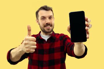 Half-length close up portrait of young man in shirt on yellow background. The human emotions, facial expression concept. Trendy colors. Negative space. Showing a smartphone and a sign of OK.