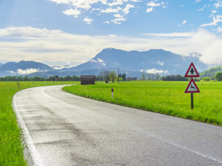 Wet asphalt road after rain at the countryside with road signs, green grass around and mountains on the horizon.