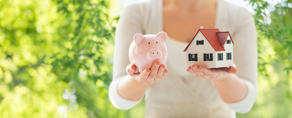 Wall Mural - mortgage, investment, real estate and property concept - close up of woman holding home or house model and piggy bank over green natural background