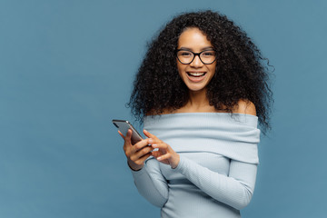 Lovely teenage girl enjoys online communication, holds modern cell phone, checks email box, connected to wireless internet, cannot imagine life without technologies, isolated on blue studio wall