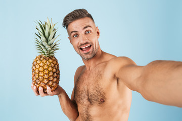 Sticker - Excited happy adult man in swimwear posing isolated over blue wall background take a selfie by camera with pineapple.