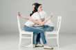 © LIGHTFIELD STUDIOS - side view of young couple embracing while sitting on white chairs and using smartphones on grey background