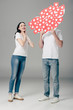 © LIGHTFIELD STUDIOS - young man hiding face behind red paper cut cards with hearts symbol near excited girl on grey background