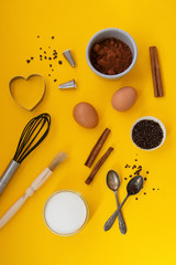 Ingredients for baking with baking utensile on yellow background - flour, wooden spoon, rolling pin, eggs, coccoa, sprinkles, banana. Top view, copy space.