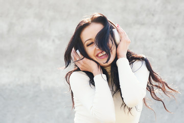 Street dancer on sunny day. Young happy sportive woman in urban background listening music in white wireless headphones and dancing. Hair fluttering in the wind 