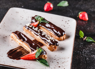 Wall Mural - Homemade cake eclairs or profiteroles with custard, chocolate and strawberries on dark background served with cup of coffee. Traditional french eclairs, dessert. Closeup