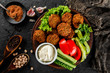 Roasted chickpeas falafel patties with garlic yogurt sauce, served with lettuce and fresh vegetables in a plate over dark stone background. Healthy vegan food, clean eating, dieting, top view