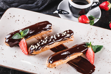 Wall Mural - Homemade cake eclairs or profiteroles with custard, chocolate and strawberries on dark background served with cup of coffee. Traditional french eclairs, dessert. Closeup