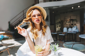 Wall Mural - Good-looking girl playing with her curly hair resting at the table with little flower vase on it. Indoor portrait of cute blonde young woman drink cocktail in cafe on blur background.