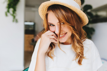 Wonderful pleased young woman smiling and playfully looking to camera. Indoor close-up portrait of attractive fair-haired female model in retro straw hat posing on blur background.