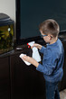Chłopiec podczas sprzątania pokoju. Nanosi detergent na mebel za pomocą aplikatora. W drugiej ręce trzyma białą ściereczkę do wycierania.